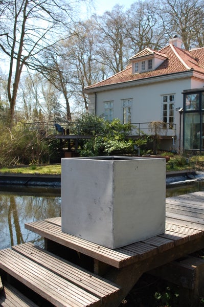 Quadratischer Pflanzkübel in Betonoptik auf einem Holzsteg an einem Teich im Garten vor einem Haus.