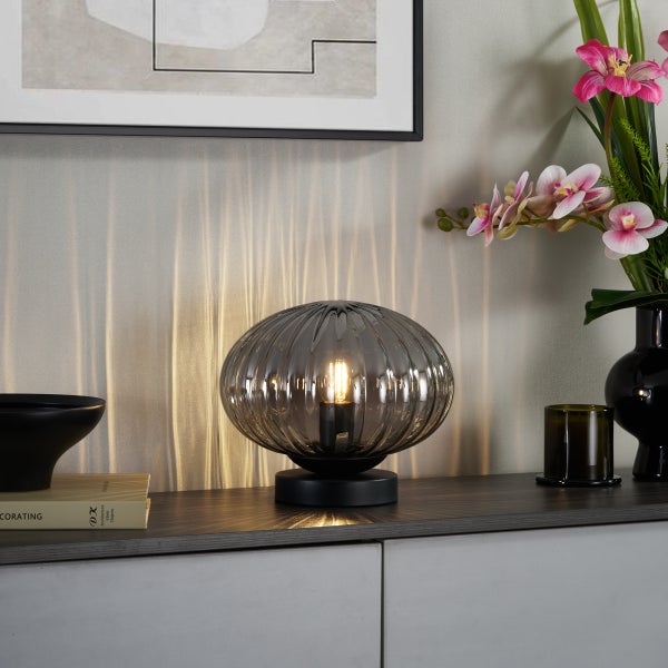 Moderne Tischleuchte mit dunklem, geripptem Glasschirm auf einem schwarzen Fuß, platziert auf einem Sideboard neben Orchideen und Dekoration.