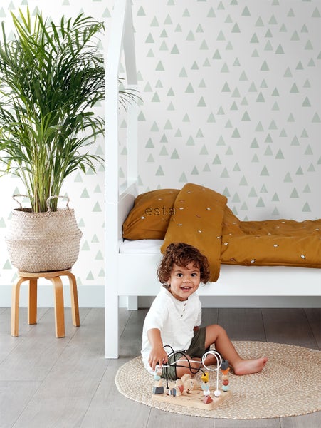 Kinderzimmer mit weißem Bett, Pflanze, gemusterten Tapeten und spielendem Kind auf einem runden Teppich