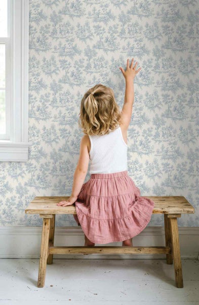 Kind sitzt auf einer Holzbank vor einer Wand mit Toile-de-Jouy-Tapete mit ländlichem Muster.