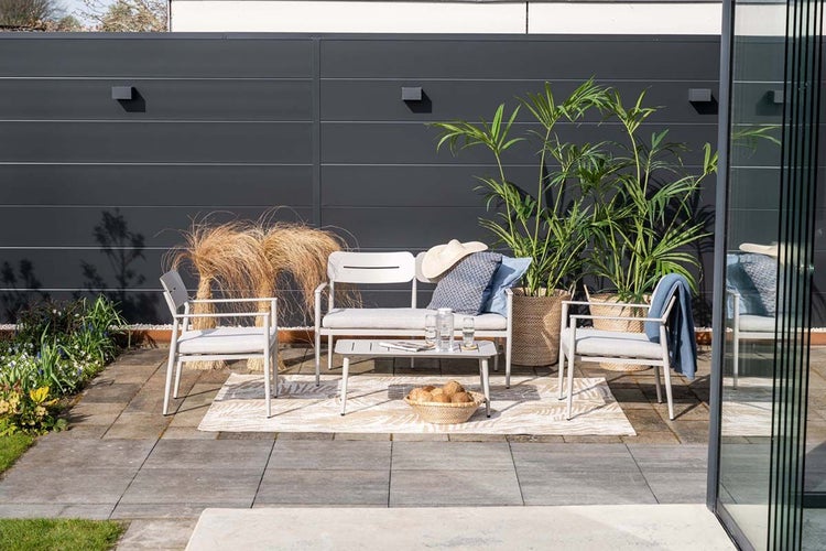 Moderne Gartenlounge aus Metall mit Sofa, zwei Sesseln und Tisch auf einer Terrasse vor einem dunklen Sichtschutzzaun.