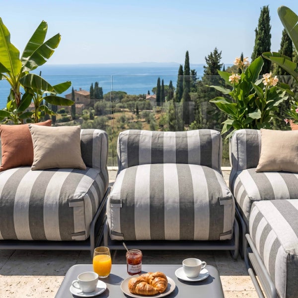 Garten-Lounge-Möbel mit grau-weiß gestreiften Kissen auf einer Terrasse mit Meerblick. Frühstück auf einem Beistelltisch im Vordergrund.