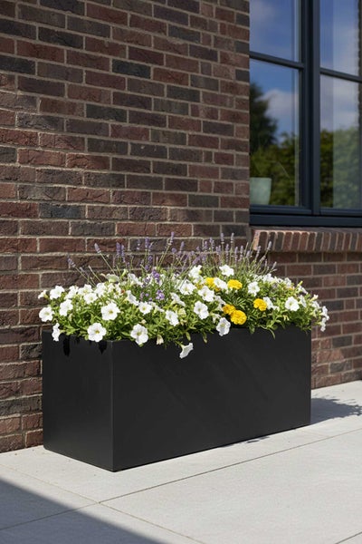 Großer rechteckiger Pflanzkasten in Schwarz vor einer Backsteinwand, bepflanzt mit weißen, gelben und violetten Blumen auf einer Terrasse.