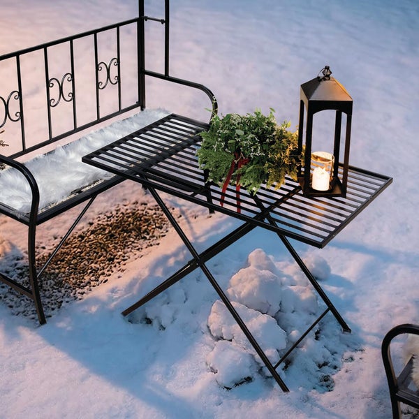 Metallgartenbank und Klapptisch im Schnee mit flauschigem Kissen, Weihnachtsgirlande und Windlicht.