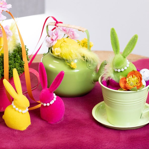 Osterdekoration mit beflockten Hasenfiguren, einer Teekanne und Metalltöpfen auf einem Tisch.
