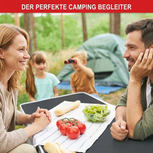 Familie beim Camping an einem Klapptisch mit Lebensmitteln, im Hintergrund ein Zelt im Wald.