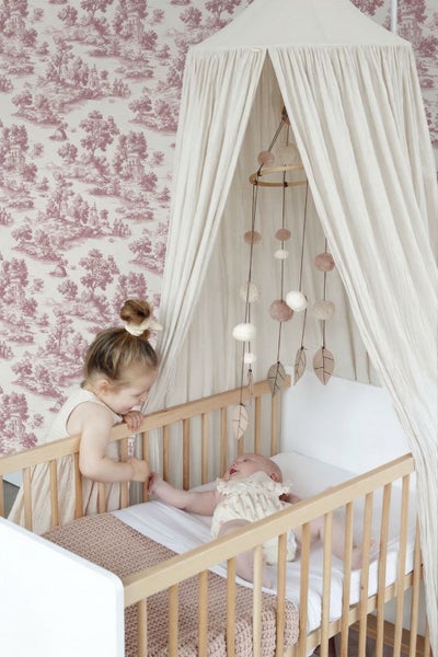 Helles Babyzimmer mit einem Gitterbett aus Holz, einem Stoffbaldachin, einem dekorativen Mobile und einer gemusterten Tapete.