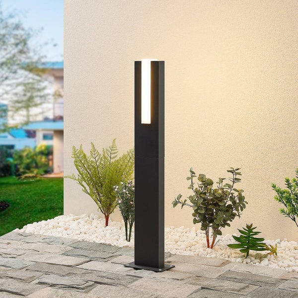 Moderne anthrazitfarbene LED-Wegeleuchte in minimalistischem Design auf einer Terrasse mit weißen Kieselsteinen und grünen Pflanzen.