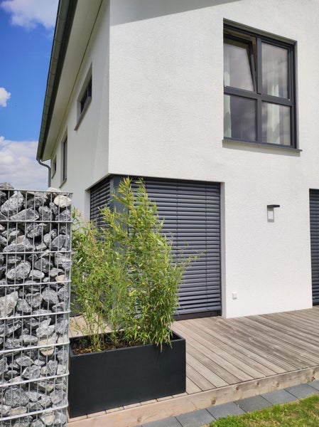 Moderne Terrasse mit Holzdeck, schwarzem Pflanzkasten mit Bambus und einer Gabionenwand aus grauen Steinen vor einer weißen Hausfassade.