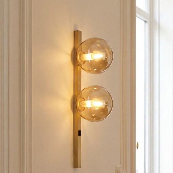 Vertikale Wandleuchte aus Messing mit zwei kugelförmigen Glasschirmen in einem hellen, klassisch gestalteten Raum.