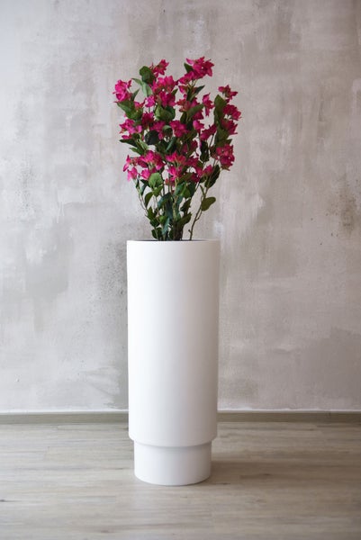 Hoher weißer zylindrischer Pflanzkübel mit einer rosa blühenden Bougainvillea vor einer strukturierten Wand.