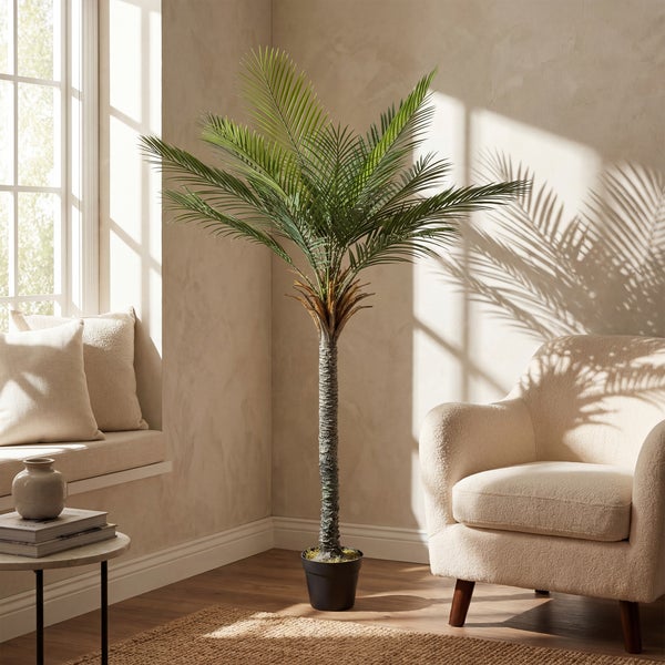 Künstliche Palme in einem schwarzen Topf, stehend in einem hellen Wohnraum neben einem beigen Sessel und einem Fenster mit Sonnenlicht.