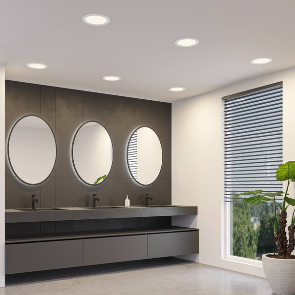 Modernes Badezimmer mit LED-Einbaustrahlern an der Decke, drei hinterleuchteten ovalen Spiegeln und dunklem Waschtisch vor einer Fliesenwand.