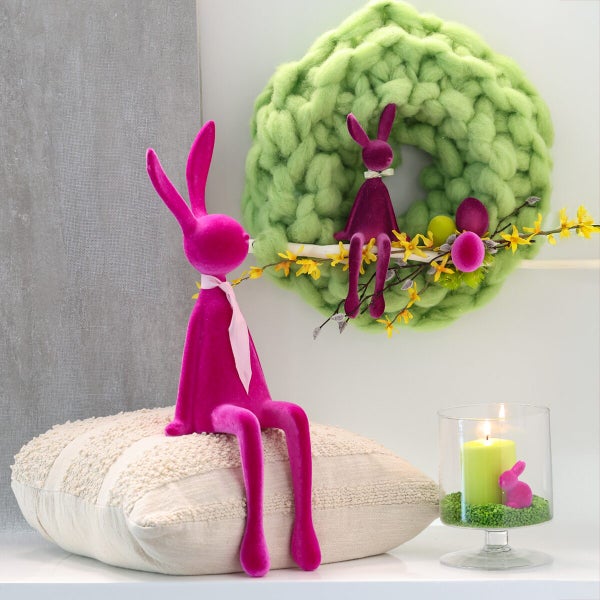 Osterdekoration mit zwei sitzenden Hasenfiguren, einem grob gestrickten Kranz, einem Kissen und einer Kerze im Glas für eine festliche Atmosphäre.