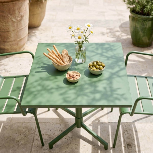 Grüner quadratischer Gartentisch aus Metall mit zwei passenden Gartenstühlen auf einer sonnigen Steinterrasse mit Snacks und Blumen dekoriert.