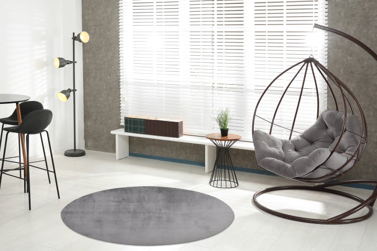 Ein moderner Wohnraum mit Hängesessel, Stehlampe, Bartisch und runden Teppich vor einem Fenster mit Jalousien und einer Sitzbank.