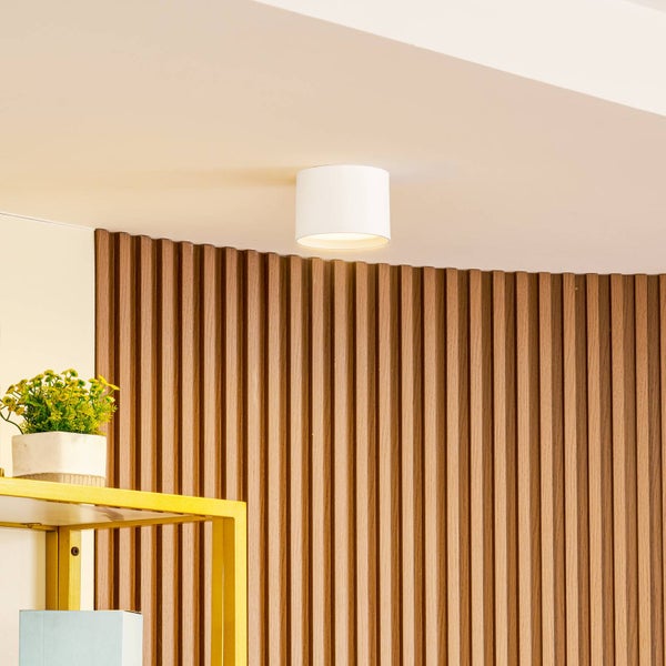 Zylindrischer Aufbauspot in Weiß an einer Decke neben einer Wand mit vertikalen Holzlamellen.