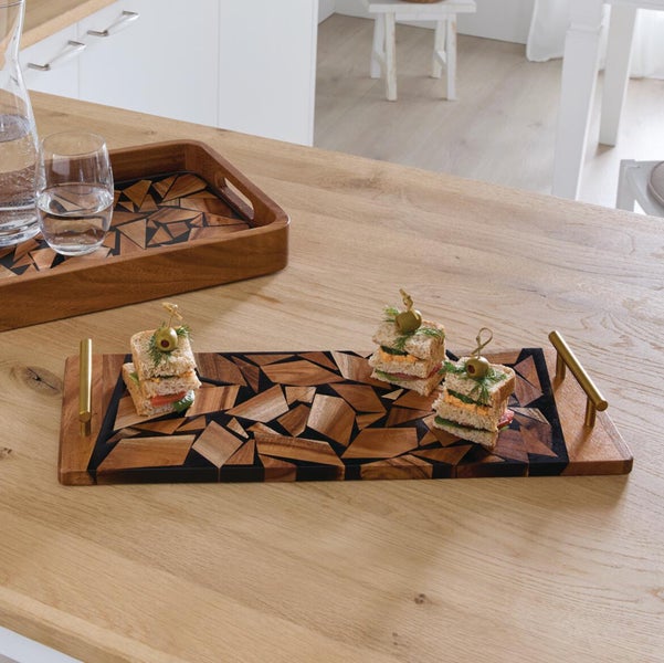 Rechteckiges Serviertablett aus Akazienholz mit geometrischem Mosaikmuster und goldfarbenen Metallgriffen auf einem hellen Holztisch.