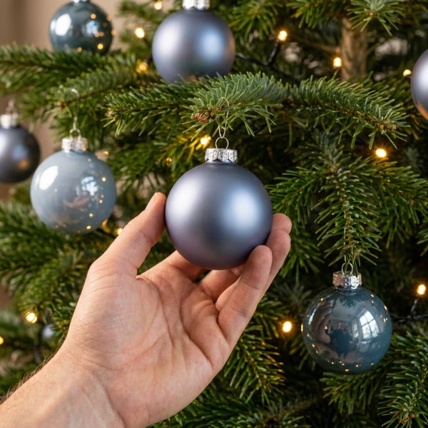 Hand hängt matte, runde Weihnachtsbaumkugel aus Glas in einen Tannenbaum mit Lichterkette.