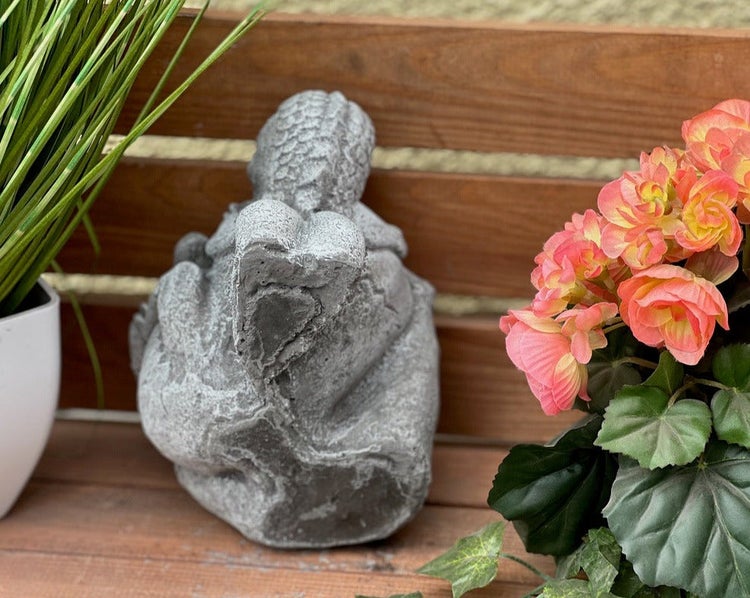 Gartenfigur aus Stein auf einer Holzbank, umgeben von Pflanzen und Blumen.
