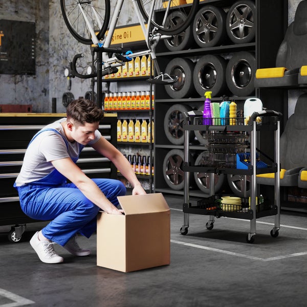 Ein Mechaniker in Arbeitskleidung kniet in einer organisierten Werkstatt vor einem Karton, neben ihm steht ein fahrbarer Werkstattwagen.