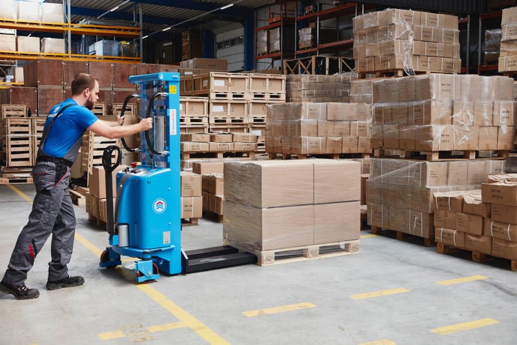 Lagerarbeiter bedient einen elektrischen Hochhubwagen zum Transport einer Palette mit Kartons in einem organisierten Hochregallager.