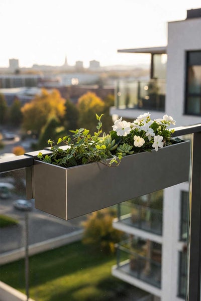 Grauer Balkonkasten aus Metall am Geländer, bepflanzt mit weißen Blumen und Efeu auf einem modernen Balkon mit Stadtblick.