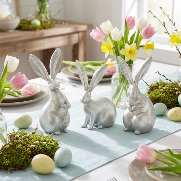 Drei stehende Osterhasen Figuren als Tischdekoration arrangiert auf einem Esstisch mit Blumen und Ostereiern als festliche Frühlingsdekoration.