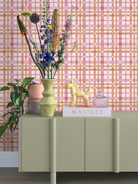 Vliestapete mit grafischem Karomuster in Rosa und Orange in einem hellen Raum hinter einem Sideboard mit Blumenstrauß und Dekoration.