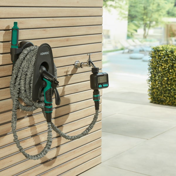 Vonroc dehnbarer Gartenschlauch auf Wandhalterung mit Spritzpistole und Bewässerungscomputer an einer Holzlattenwand im Garten.