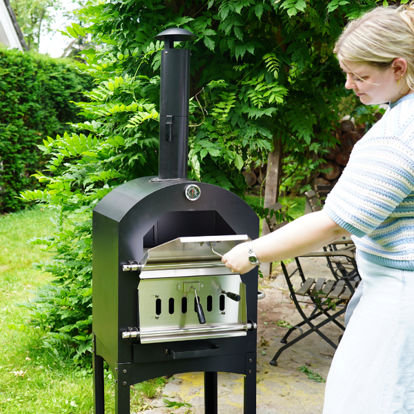 Eine Frau öffnet die Klappe eines schwarzen Outdoor-Pizzaofens mit Thermometer in einem Garten mit viel Grün.