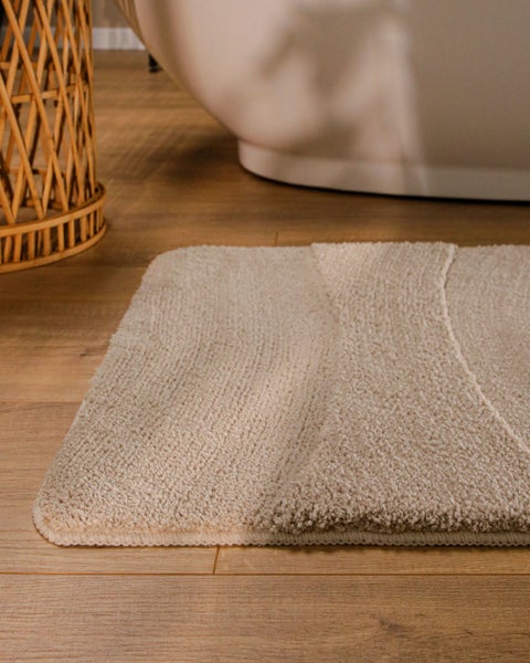 Beige Badematte mit strukturiertem Wellenmuster aus weichem Hoch-Tief-Flor auf Holzboden neben einer Badewanne.