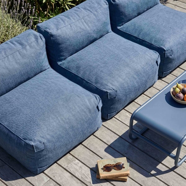 Drei blaue Outdoor-Lounge-Module aus Textilgewebe auf einer Holzterrasse neben einem Beistelltisch aus Metall mit Dekoration.