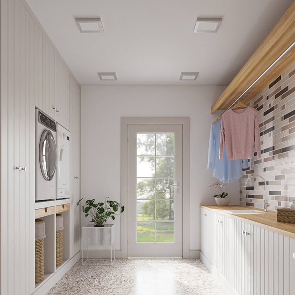 Helle Waschküche mit weißen Einbauschränken, Waschmaschine, Trockner, Holz-Arbeitsplatte mit Spüle und quadratischen LED-Panels an der Decke.