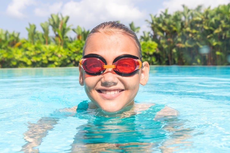 Kind mit roter Schwimmbrille in einem hellen Swimmingpool mit Palmen im Hintergrund.