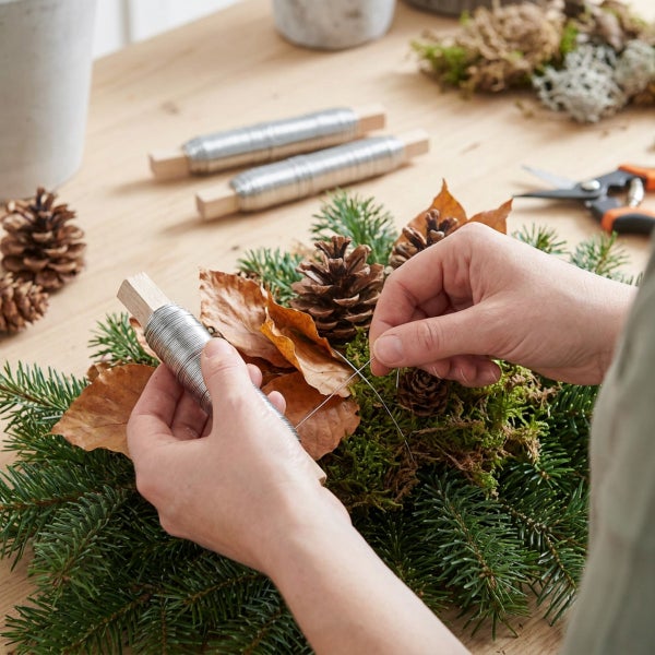 Person beim Binden eines Adventskranzes aus Tanne Moos Zapfen und Laub mit Wickeldraht auf einem Holztisch