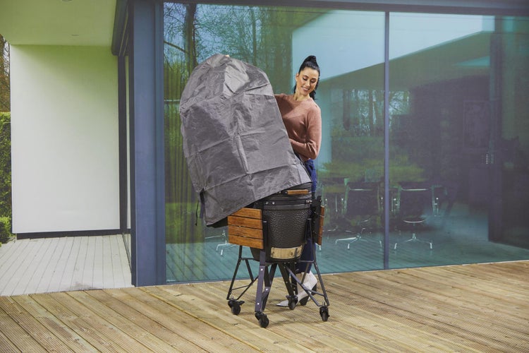 Frau legt eine graue Schutzhülle über einen Keramikgrill auf einer Terrasse