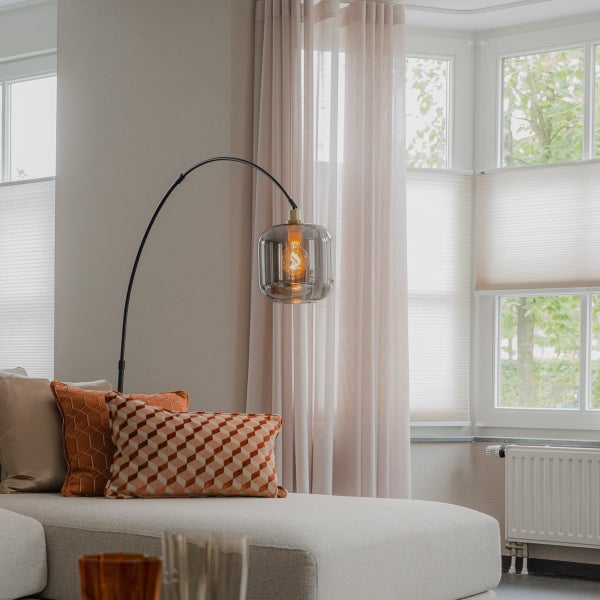 Moderne Bogenstehlampe mit schwarzem Gestell und Glasschirm aus Rauchglas neben einem Sofa in einem hellen Wohnzimmer