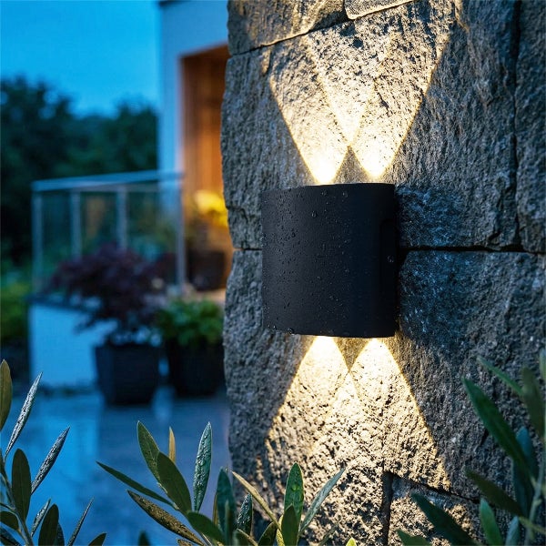 Schwarze Außenwandleuchte mit Lichtkegeln nach oben und unten an einer dunklen Steinwand.