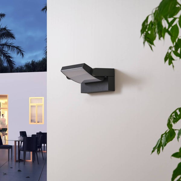 Moderne LED-Außenwandleuchte in Anthrazit an einer weißen Wand. Im Hintergrund eine organisierte Terrasse mit Sitzgruppe bei Dämmerung.