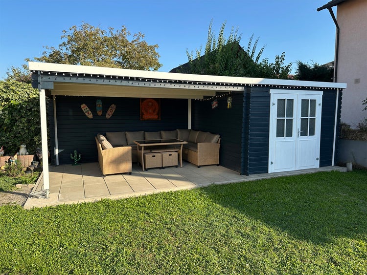 Modernes Gartenhaus in Anthrazit mit weißer Doppeltür und integrierter, überdachter Terrasse inklusive Rattan-Lounge-Set auf Steinplatten.