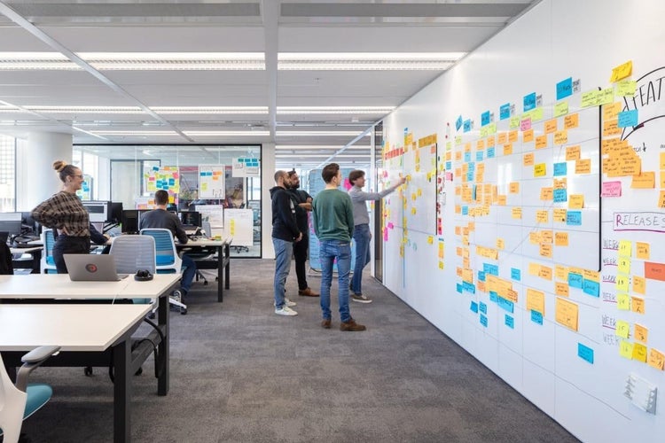 Modernes, helles Großraumbüro mit Whiteboard-Wänden, bunten Haftnotizen, Glaswänden und Mitarbeitern in organisierter, großzügiger Atmosphäre.