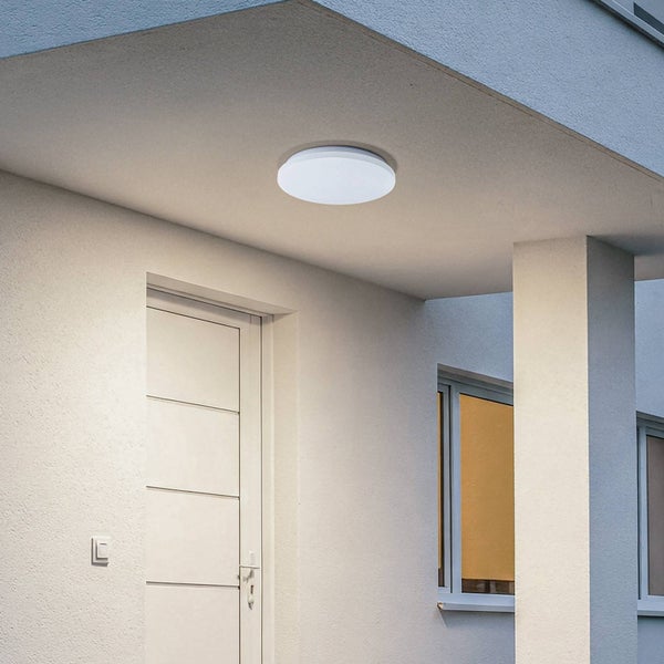 Runde LED-Außenleuchte montiert an einem Dachvorsprung im Eingangsbereich eines hellen Hauses mit weißer Tür.