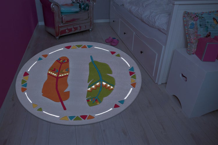 Runder Kinderteppich mit Feder Motiven in einem Kinderzimmer mit Bett und Sessel auf einem Holzboden.