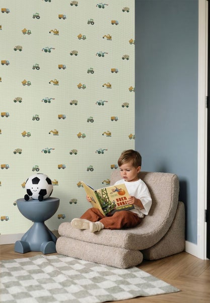 Papiertapete mit Baufahrzeugen in einem Kinderzimmer. Ein Kind sitzt in einem Sessel und liest ein Buch neben einem Beistelltisch.