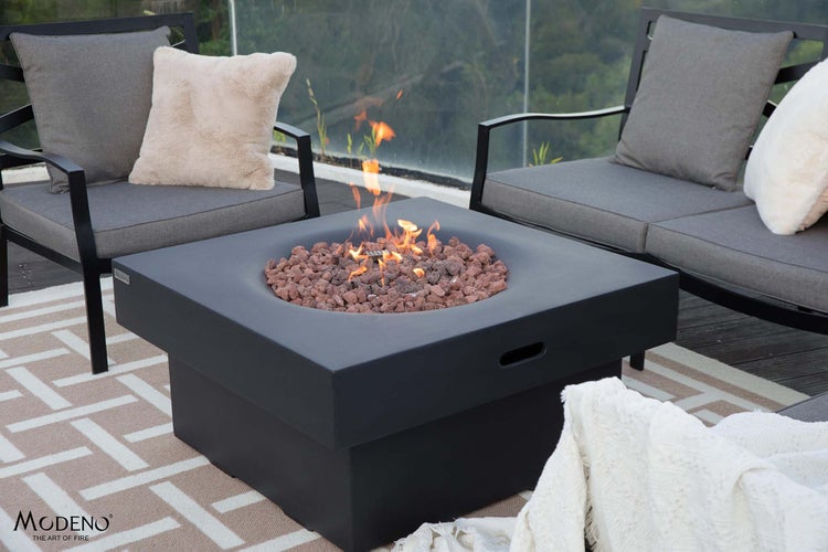 Quadratischer Gaskamin von Modeno mit Lavasteinen und Flamme auf einer Terrasse mit Gartenmöbeln und Teppich.