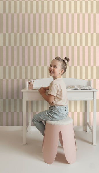 Ein lächelndes Kind sitzt an einem weißen Schreibtisch vor einer Tapete mit geometrischem Streifenmuster in Beige und Rosa.