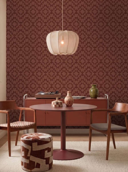 Wohnzimmer mit gemusterter Tapete, rundem Esstisch, Holzstühlen, einem Pouf und einer Hängeleuchte im retro Stil.