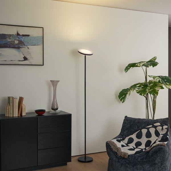 Moderne Stehleuchte mit schlankem schwarzen Standfuß und rundem flachen Leuchtenkopf in einem hellen Wohnzimmer mit Sideboard und Sessel.