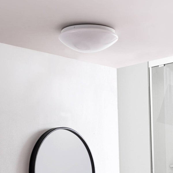 Weiße runde Deckenleuchte in einem hellen Badezimmer über einem runden schwarzen Spiegel und einer Glasdusche.